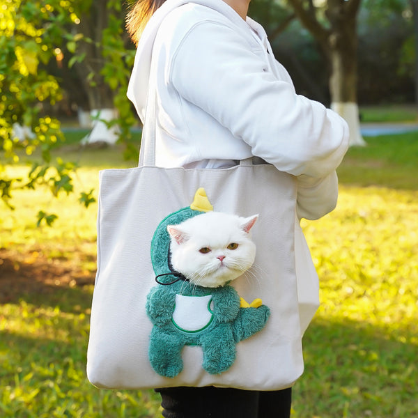Person holding a tote bag with a cat in a dinosaur costume on a grassy outdoor setting