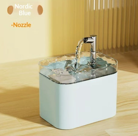 A Nordic Blue water filter pitcher with a chrome faucet spout dispensing water, set on a light wood surface against a warm beige background.