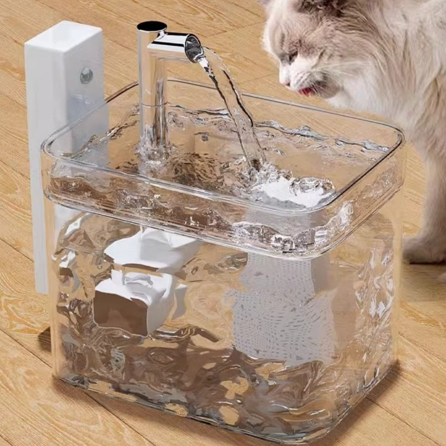 Clear pet water fountain with a cat drinking from it on a wooden floor.