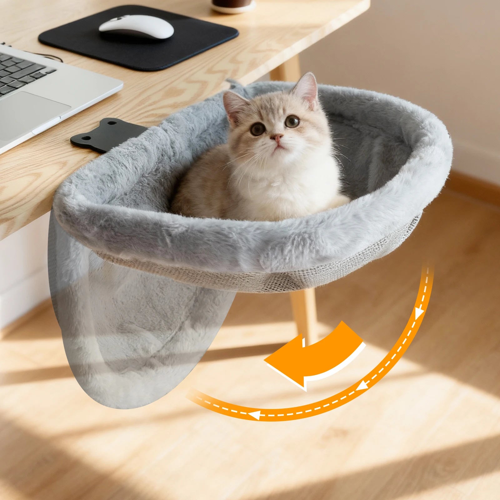 Cat sitting in a gray pet hammock on a wooden desk with a laptop and mouse.