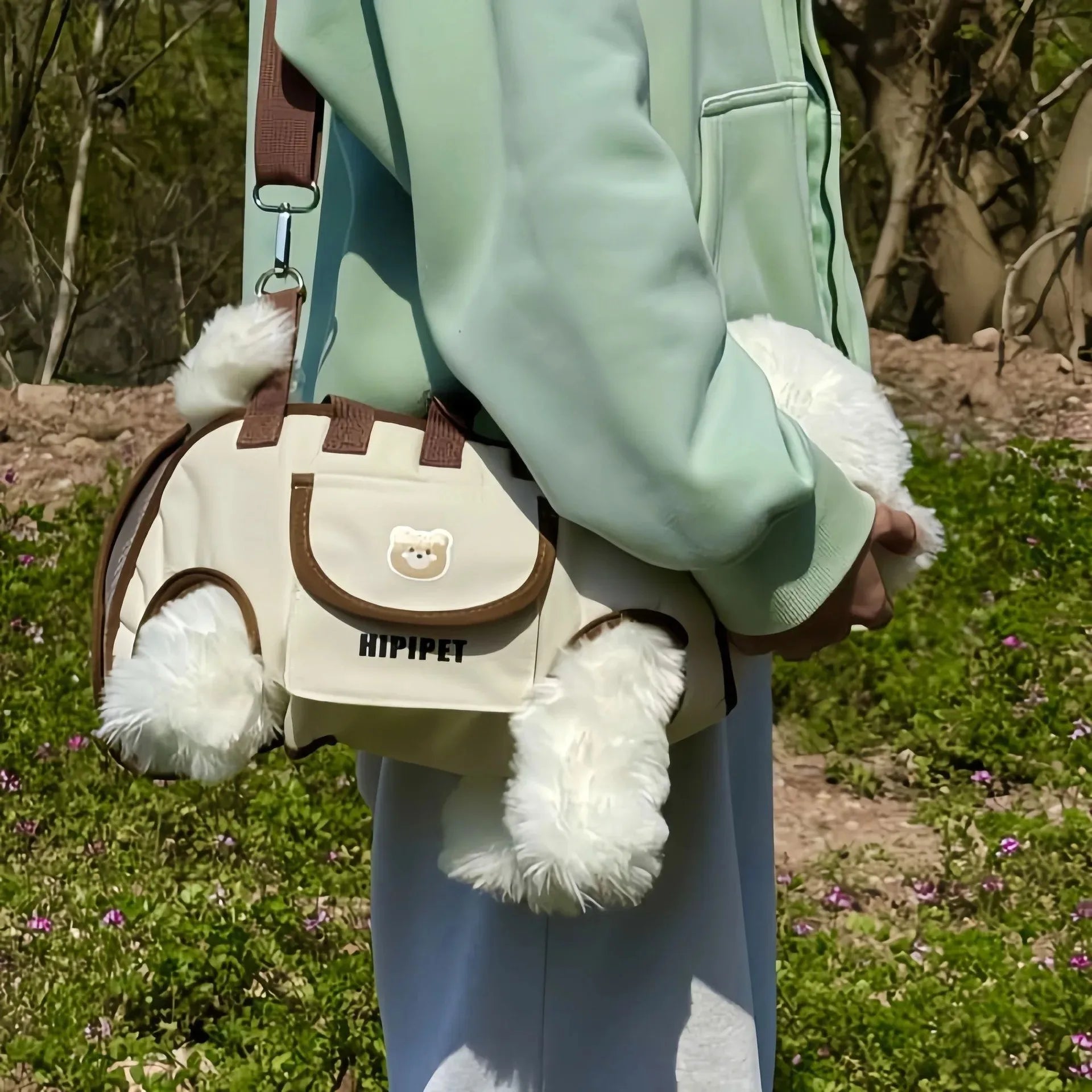 Person holding a beige bag with brown accents and white fluffy ends, standing in a natural setting.